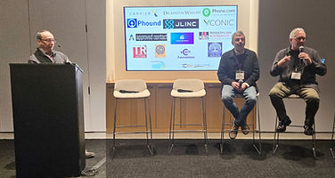 Three people on a stage, one at a podium, in front of a screen of sponsor logos