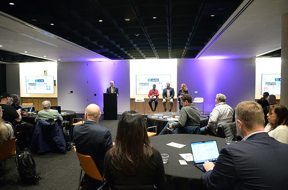 A wide view of a purple-lit room with a panel on stage and attendees at tables