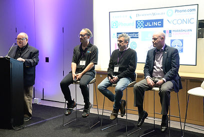 A podium and three seated panelists in front of a screen of sponsor logos