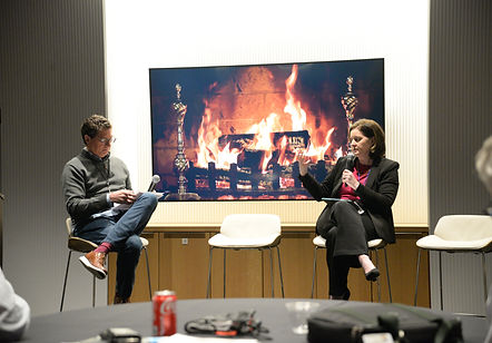 Two attendees seated on stools in front of a screen showing a fireplace